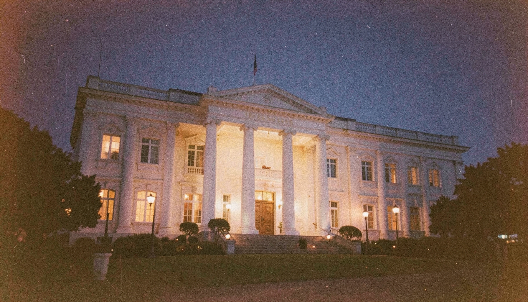 A grainy analog film photograph of the Presidential Villa at twilight, white columns glowing under soft exterior lights, deep indigo sky, stately and calm.