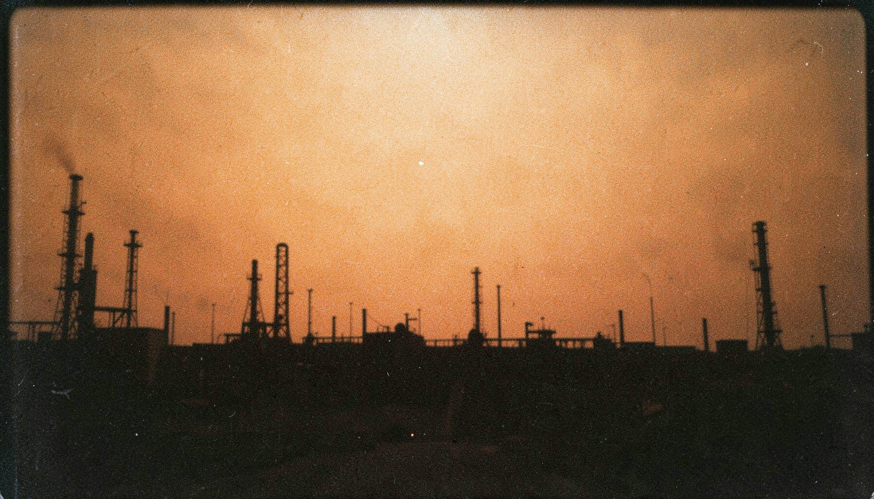 A grainy analog film photograph of oil infrastructure silhouetted against a hazy sunset, rust-orange sky, dark industrial shapes, warm amber and charcoal tones.