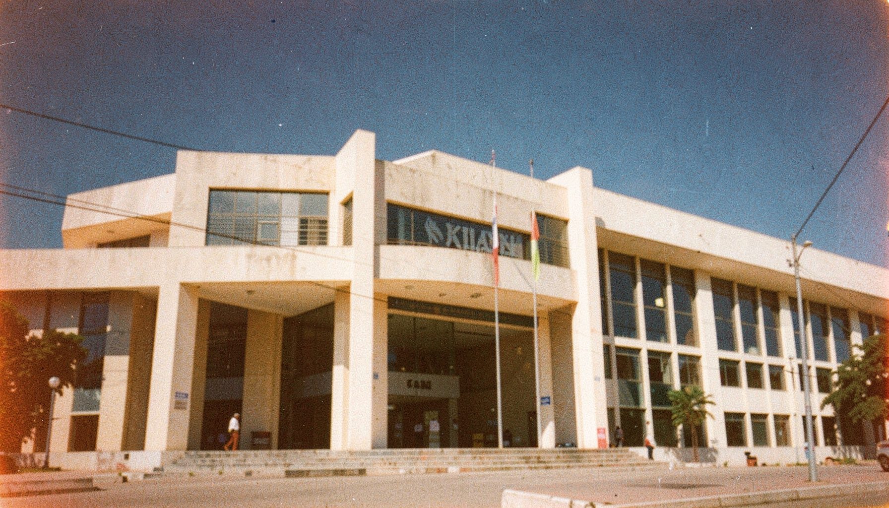 A grainy analog film photograph of a government building facade in Abuja, pale beige concrete under harsh midday sun, deep blue sky, muted institutional tones.